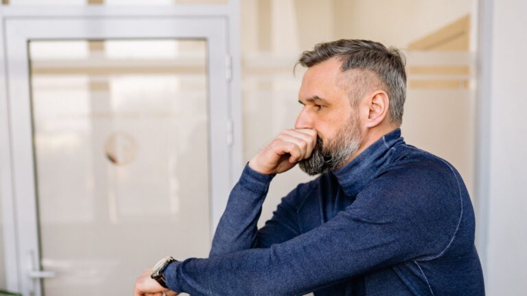 A contemplative bearded man in blue sweater leans on a chair